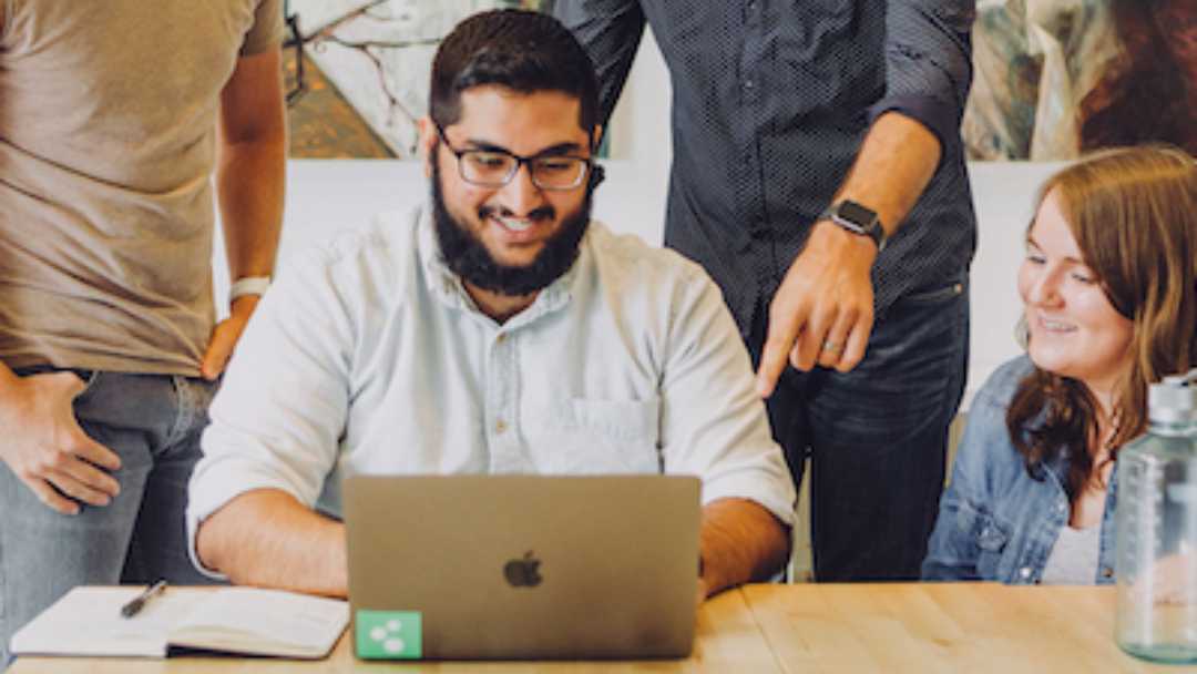 Man sitting at his laptop on a desk with people behind him