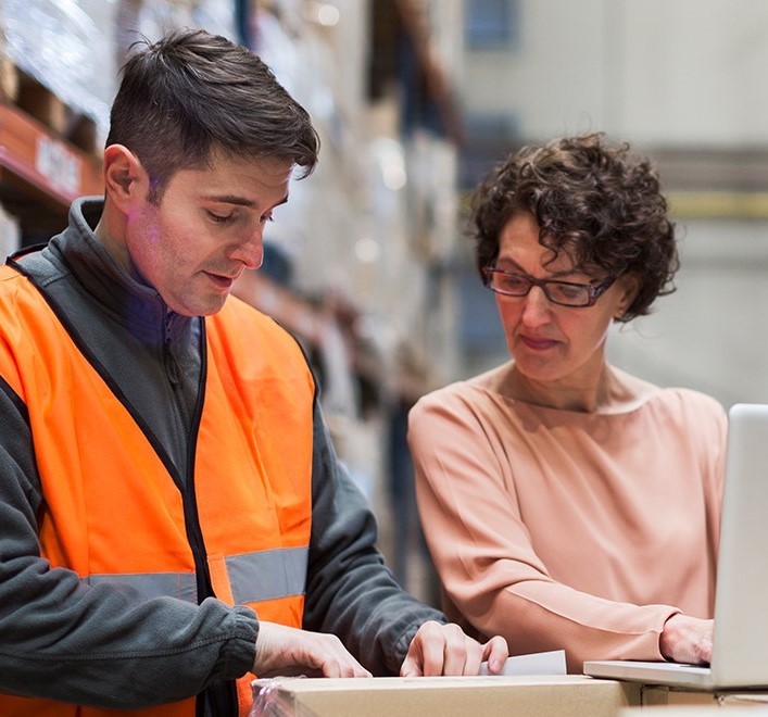 warehouse worker walking to woman in business attire while looking at a clipboard 