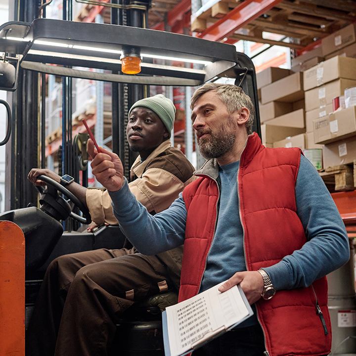 a man driving a sit down forklift with another man in a red vest pointing somewhere else in the warehouse holding a clipboard